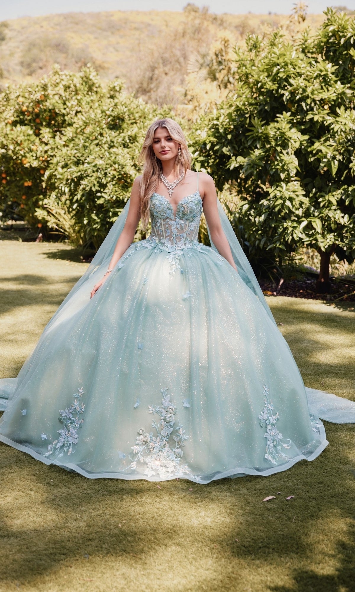 A woman stands outdoors in a garden wearing the Sage Green Quinceanera Dress: Juliet JT1466H, featuring floral embroidery, a sheer corset bodice, and a glittering, voluminous skirt.
